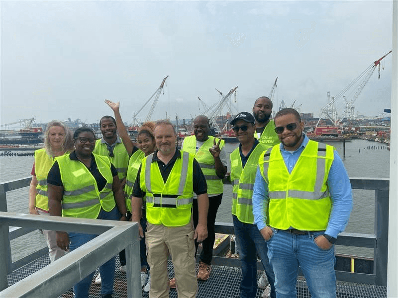 A site visit team in safety vests at a port rail terminal, with cranes and container yards behind them